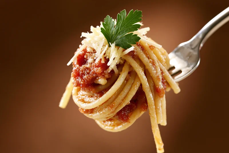Fork twirling spaghetti with tomato sauce, grated cheese and parsley against brown background