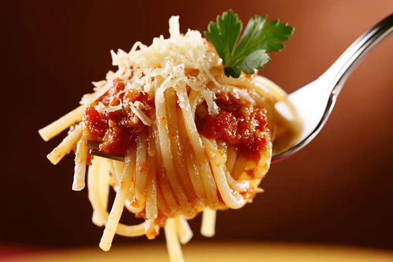 Fork twirling spaghetti with tomato sauce and grated cheese, garnished with parsley, against a warm brown backdrop