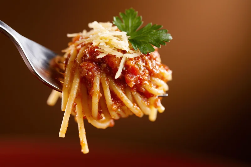 Fork holding spaghetti with tomato sauce, cheese and parsley against warm background