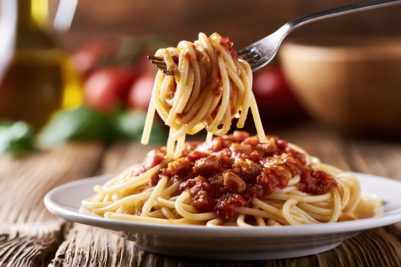 Fork twirling spaghetti with tomato sauce and pancetta above a plate on a wooden table