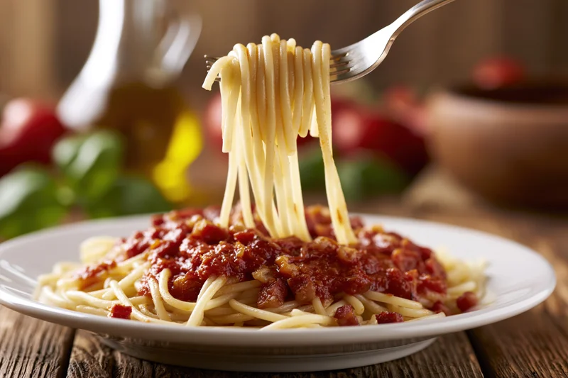 Fork twirling spaghetti with tomato sauce and pancetta lifted from a white plate on a wooden table
