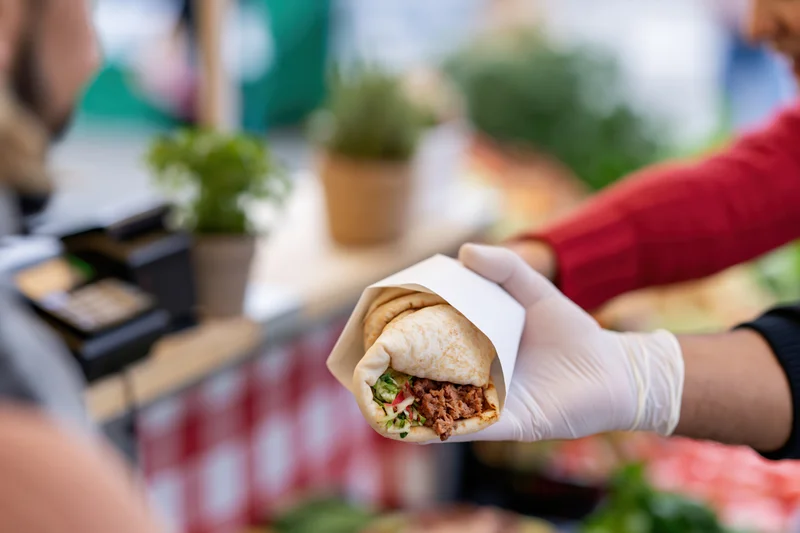 Gloved hand holding a stuffed flatbread wrap extended toward a customer at an outdoor food stand