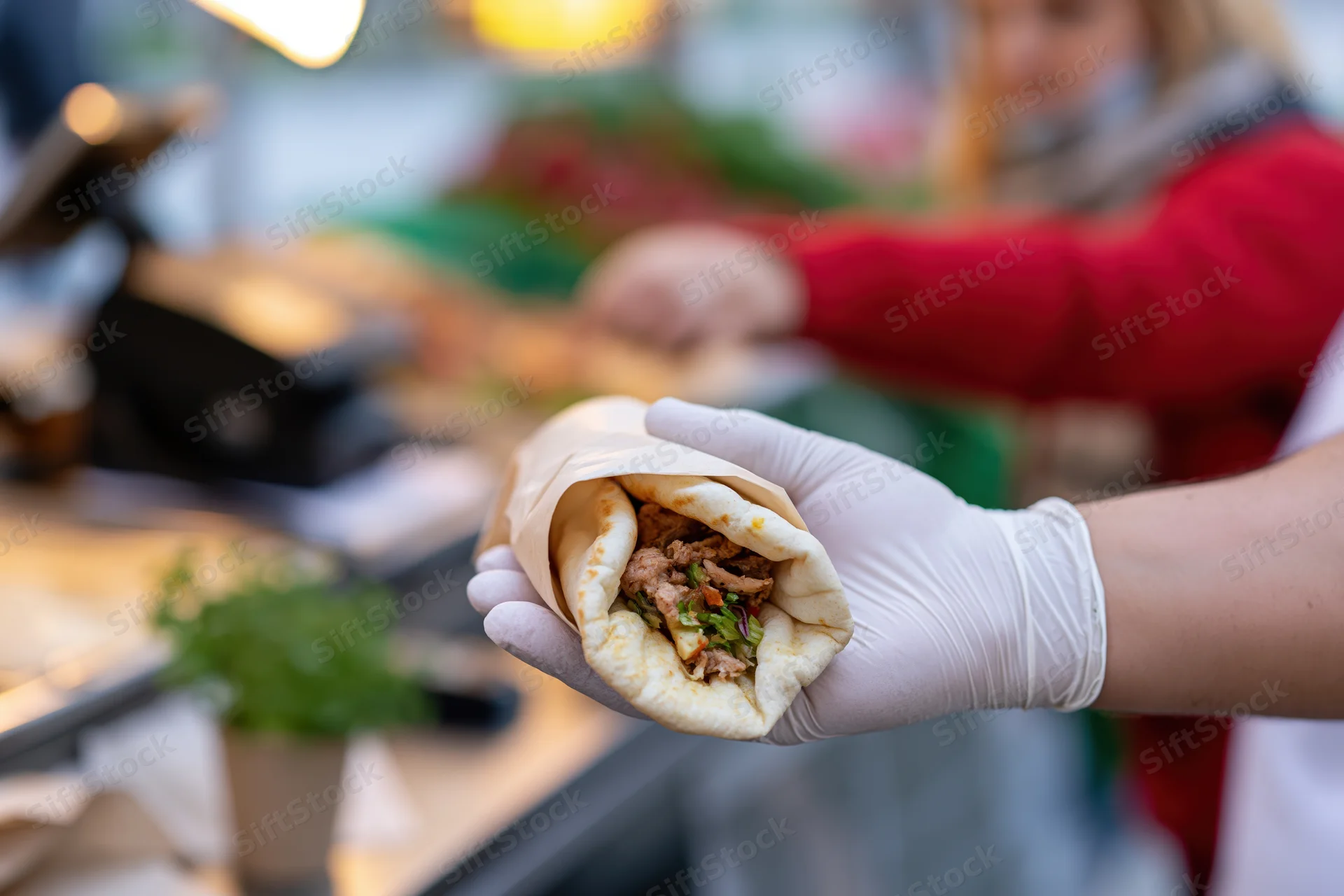 Gloved hand holding a meat and vegetable stuffed flatbread wrap extended toward a customer at a food stall