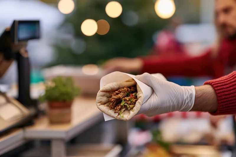 Gloved hand holding a meat and vegetable filled flatbread wrap extended toward a customer at a restaurant counter