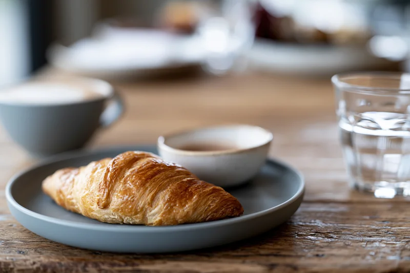 Golden-brown croissant on a grey ceramic plate with coffee and water on a wooden table
