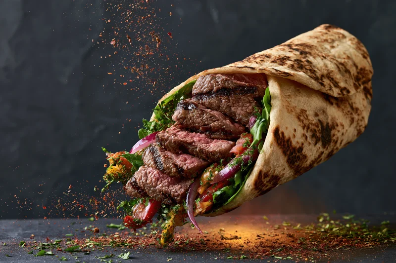 Grilled beef wrap mid-air with sliced steak and fresh greens bursting from tortilla on dark surface