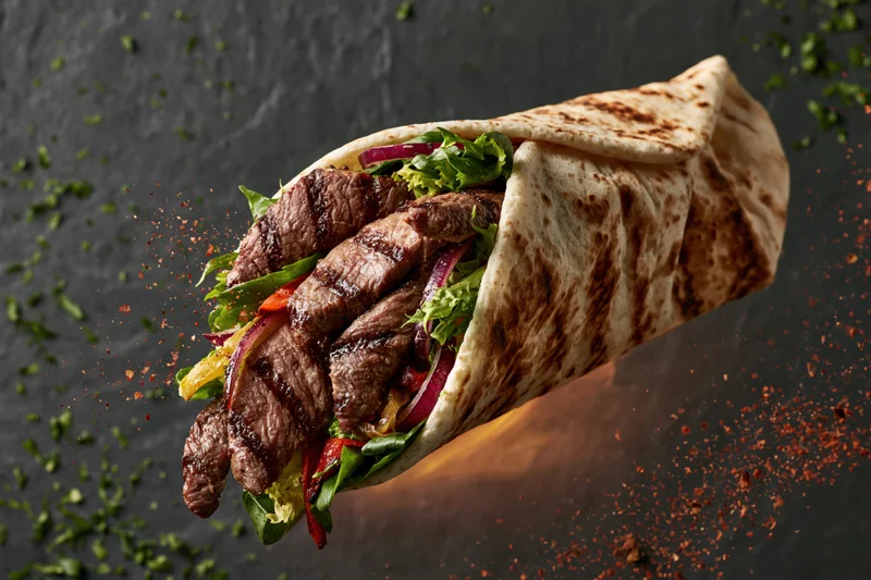 Grilled beef wrap mid-air with sliced steak and fresh greens on dark background