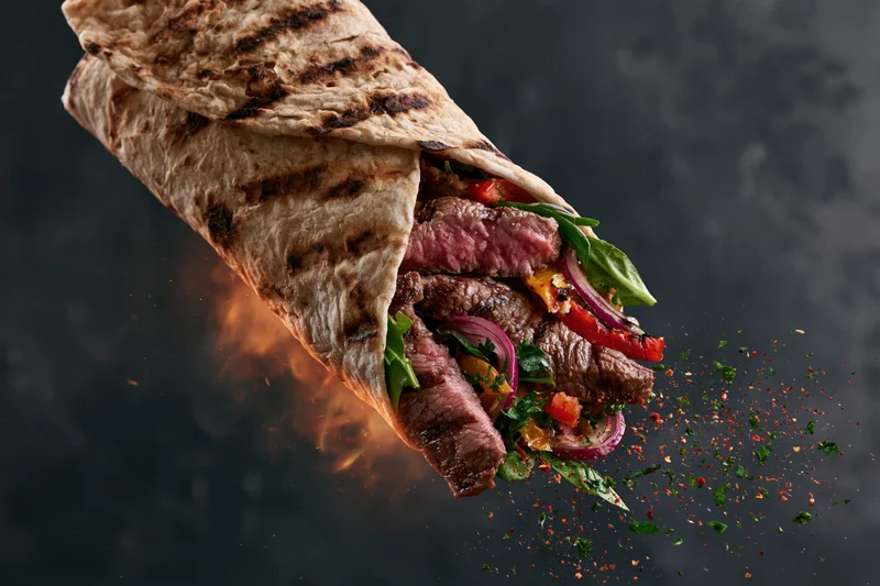 Grilled beef wrap mid-air with sliced steak and fresh greens bursting from the tortilla against a dark background