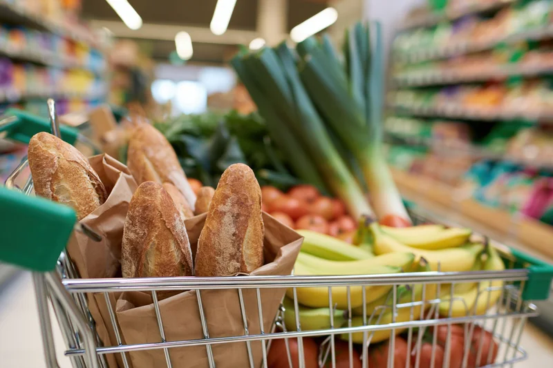 Shopping cart filled with fresh vegetables, bread, and fruit in a supermarket aisle