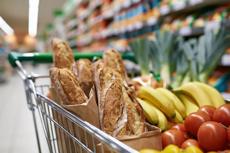 Shopping cart filled with fresh vegetables and bread in a supermarket aisle