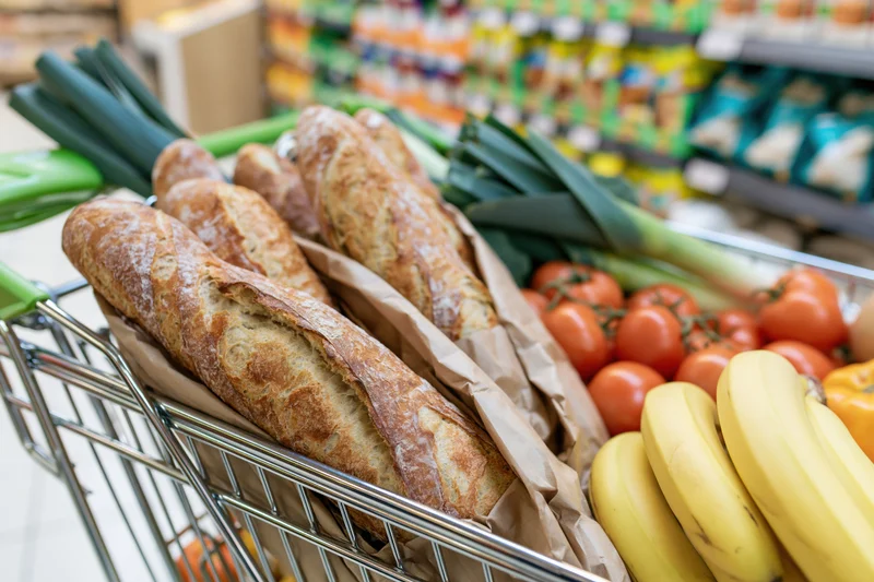 Shopping cart filled with fresh vegetables, bread, and bananas in a supermarket aisle