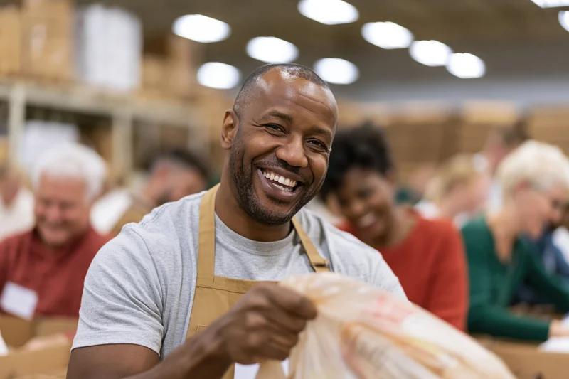 Smiling african man in apron packing food donations in warehouse with volunteers