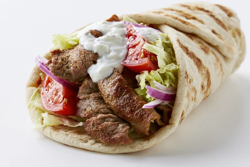 Gyro wrap filled with grilled meat, shredded lettuce, tomato, red onion and white sauce on white background