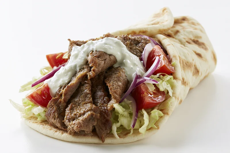 Gyro wrap filled with seasoned meat, shredded lettuce, tomato, red onion and creamy sauce on white background