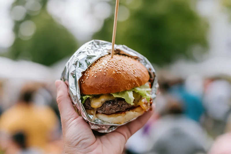 Hand holding a large gourmet burger wrapped in foil outdoors