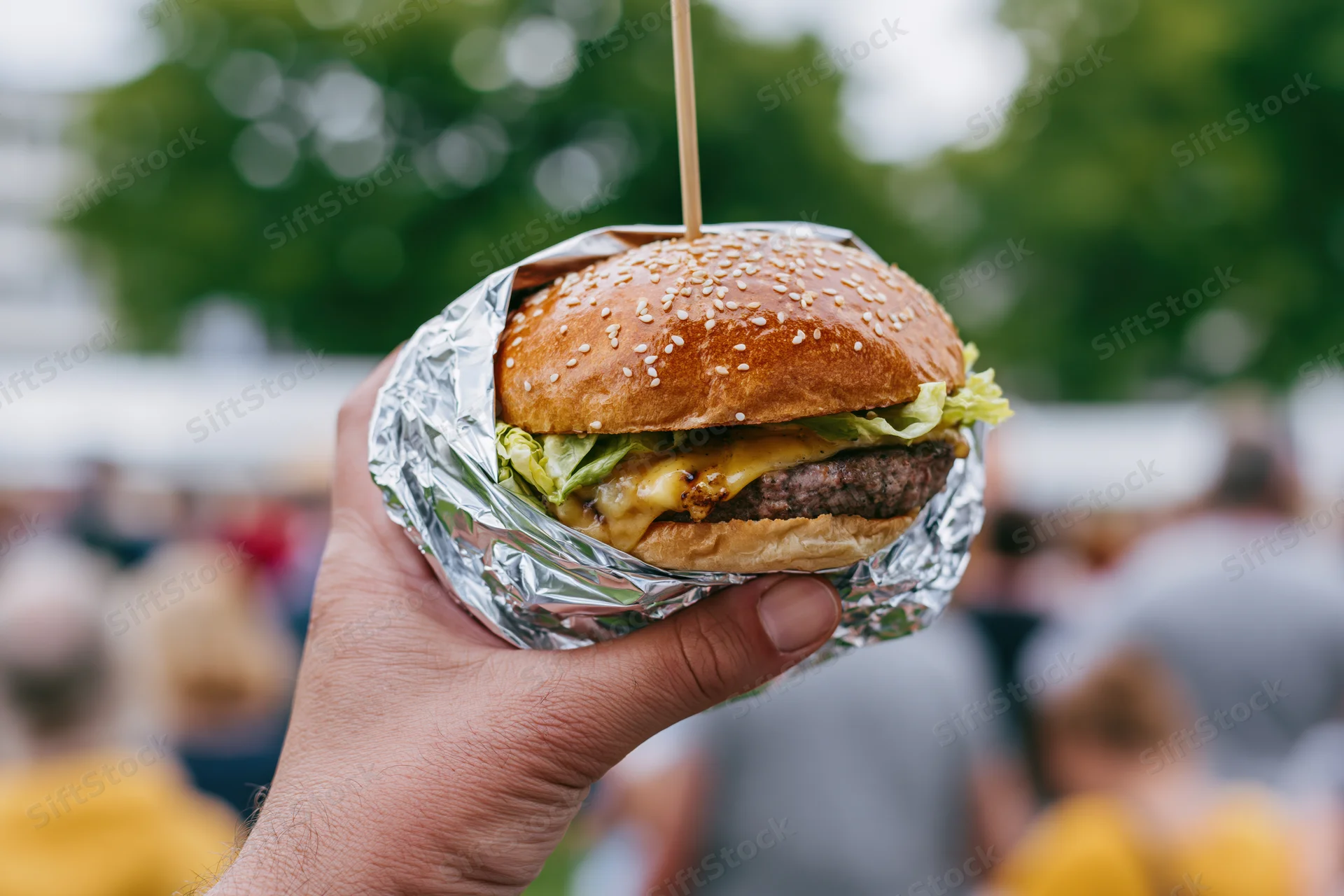 Hand holding a cheeseburger wrapped in foil at an outdoor festival