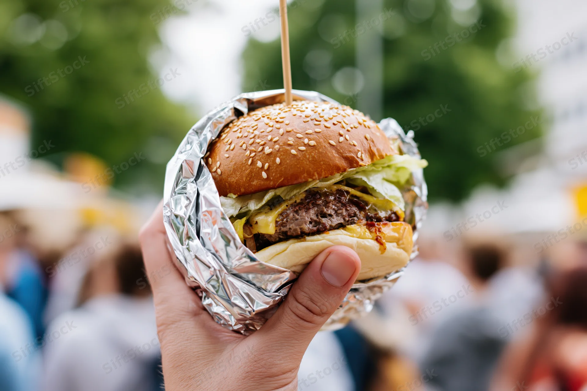 Hand holding a foil-wrapped burger with cheese and sesame seeds outdoors at a festival