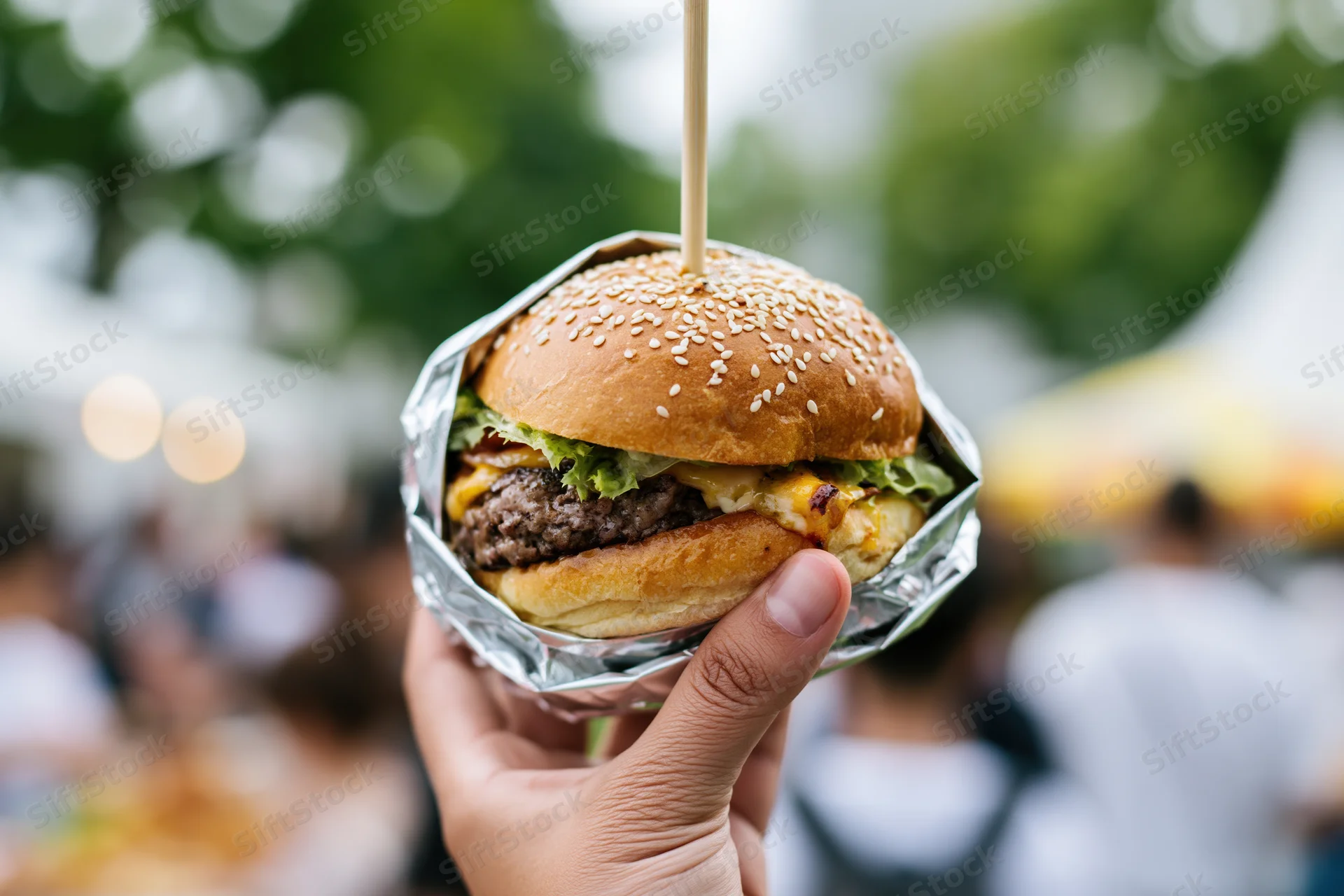 Hand holding a large gourmet burger wrapped in foil at an outdoor event