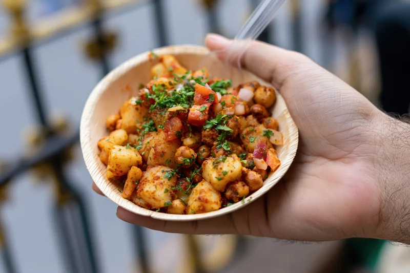 Hand holding a paper leaf bowl filled with spiced Indian street food of potatoes and chickpeas garnished with cilantro and tomato