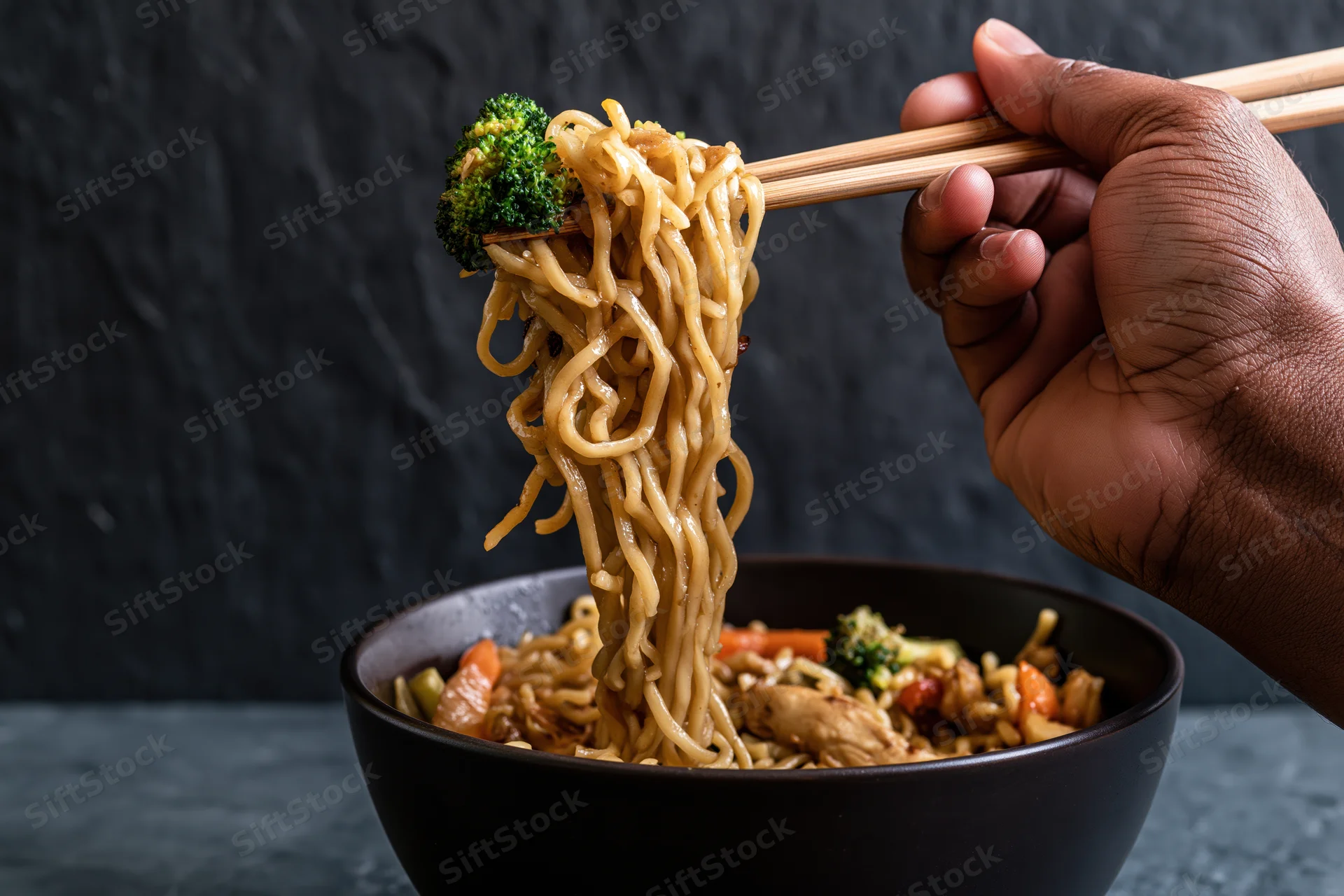 Hand lifting noodles with chopsticks from a bowl of stir-fry with vegetables and chicken