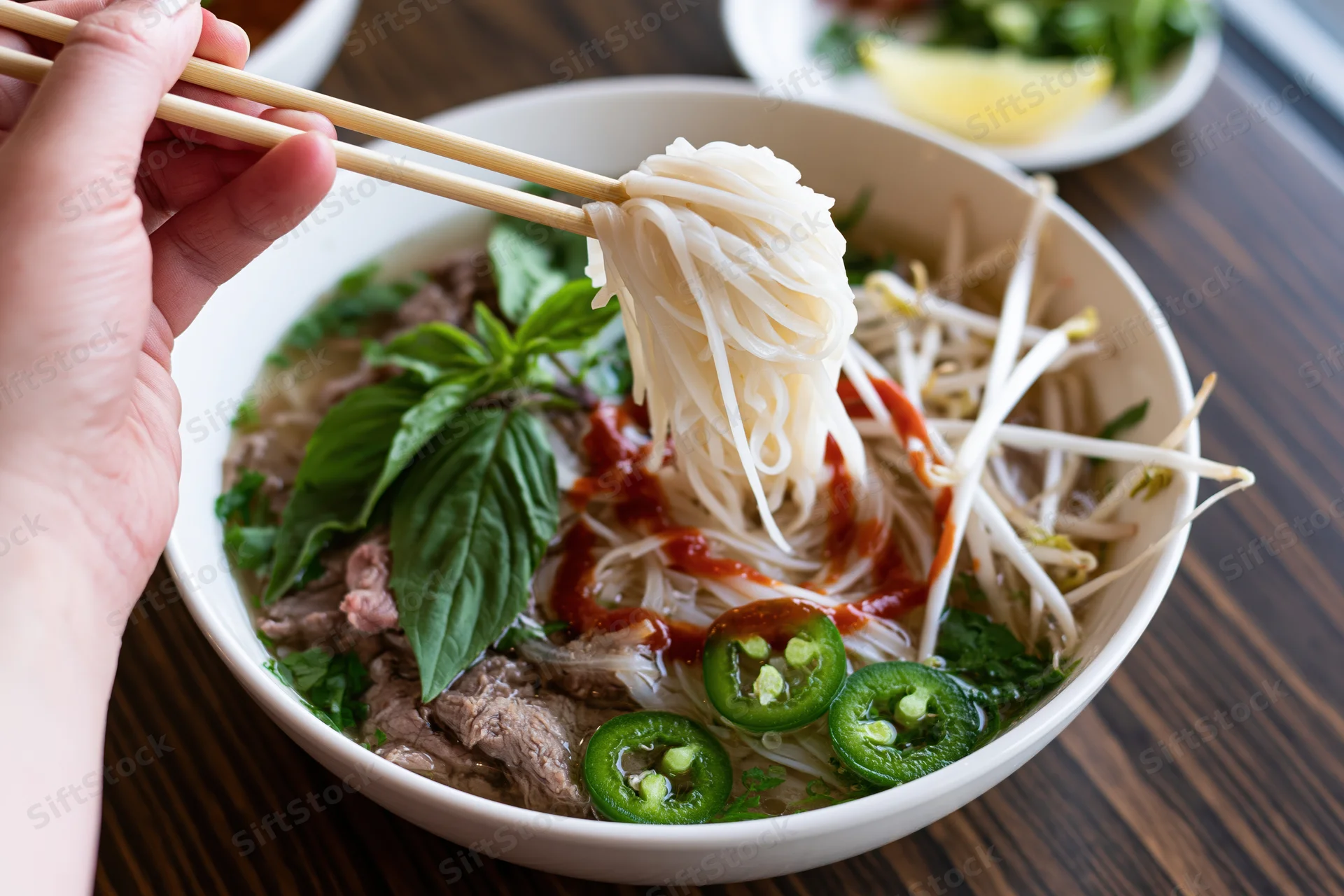 Hand lifting noodles with chopsticks from a bowl of pho on a wooden table