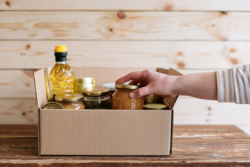 Hand reaching into open cardboard box filled with glass jars and cans on wooden table
