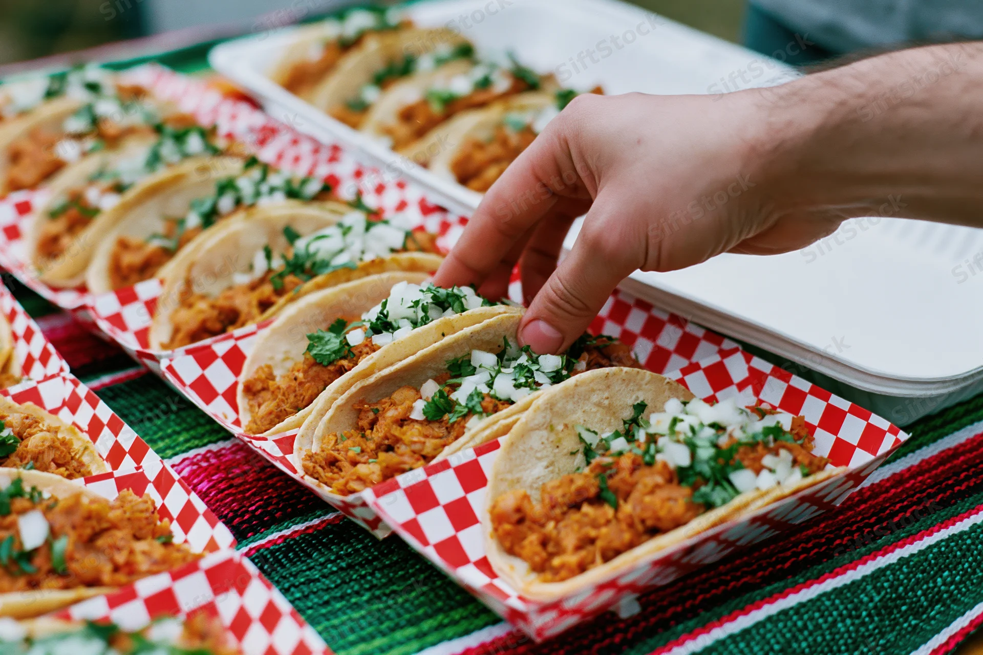 Hand reaching to pick up a street taco from a red-checkered paper tray on a striped blanket