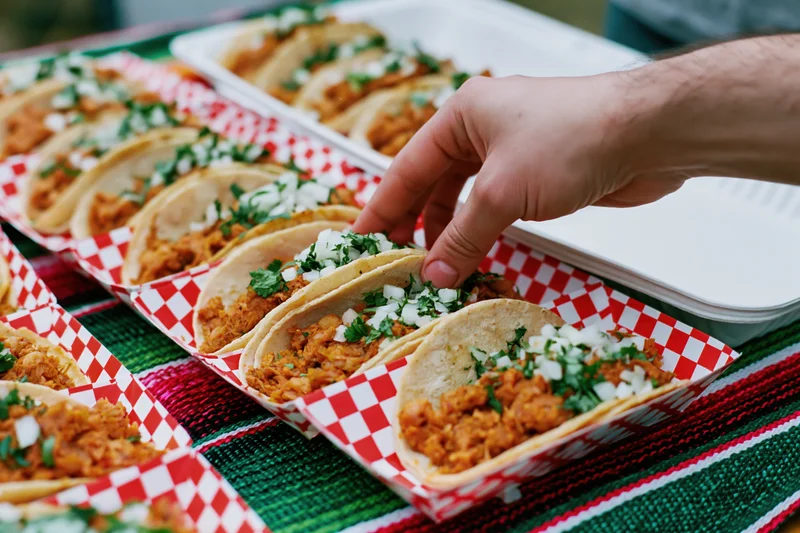 Hand reaching to pick up a street taco from a red-checkered paper tray on a striped blanket