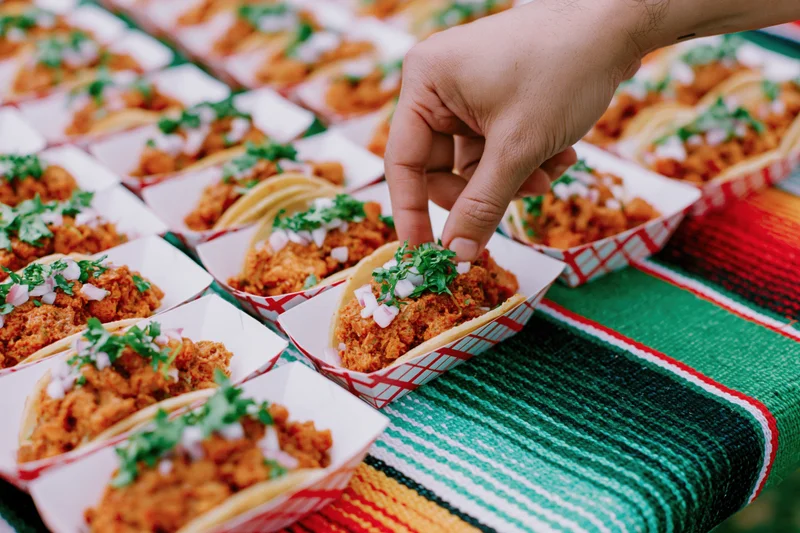 Hand reaching to pick up a street taco from a table covered with a colorful striped blanket at an outdoor event