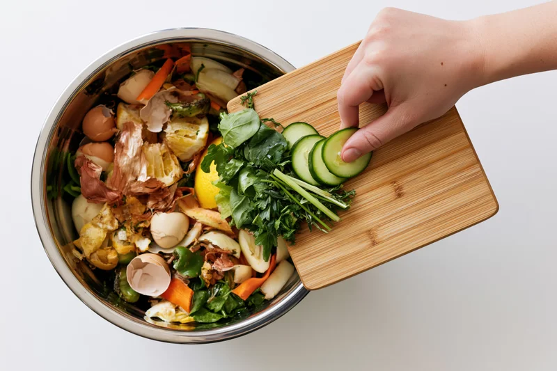Hand scraping vegetable scraps from wooden cutting board into metal compost bowl