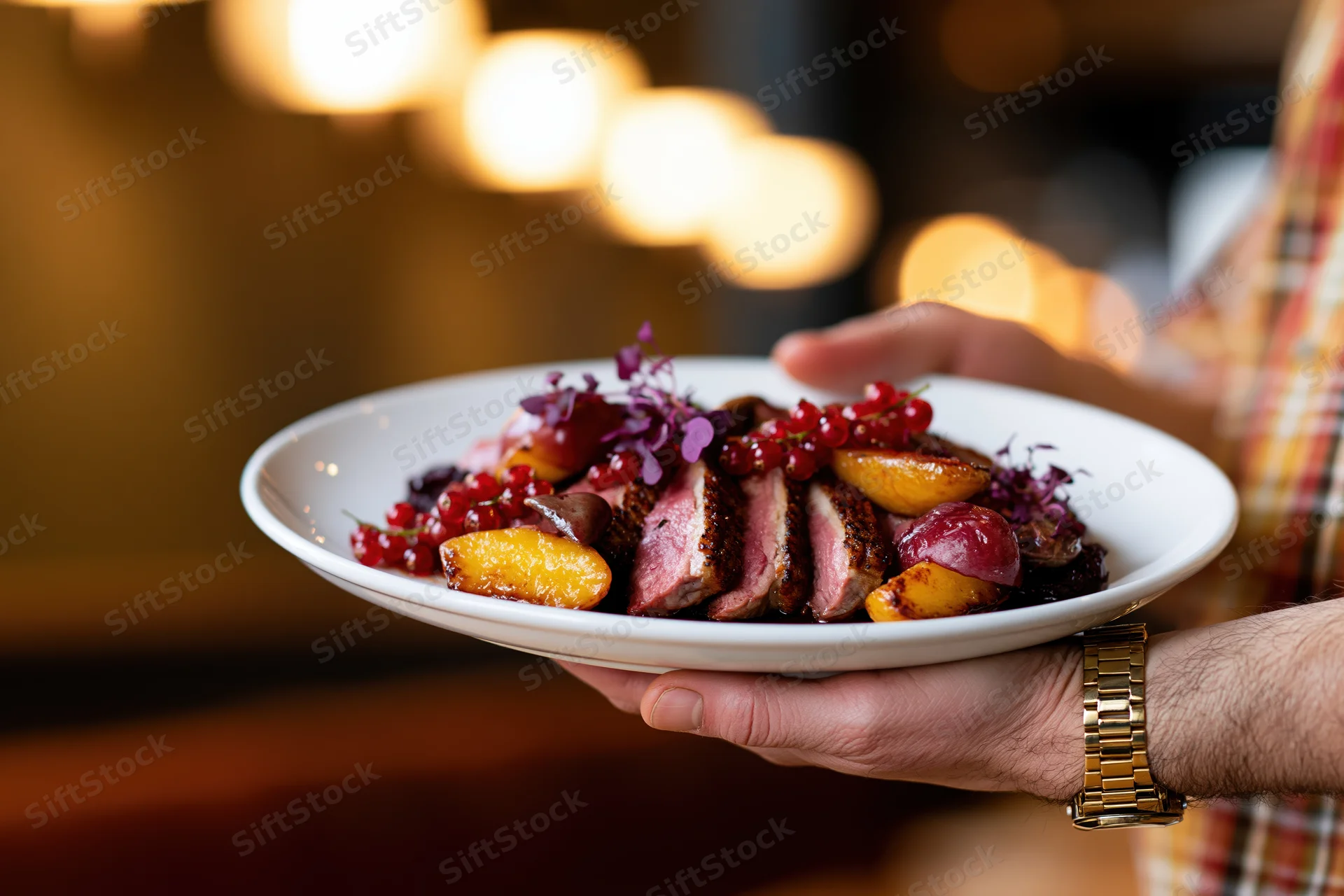 Hand holding a plated seared duck dish with roasted fruit and garnish in a restaurant setting