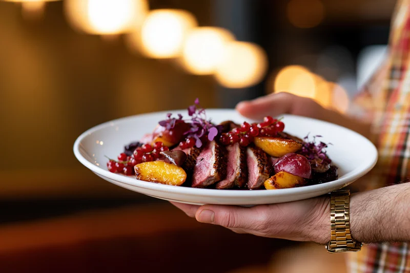 Hand holding a plated seared duck dish with roasted fruit and garnish in a restaurant setting