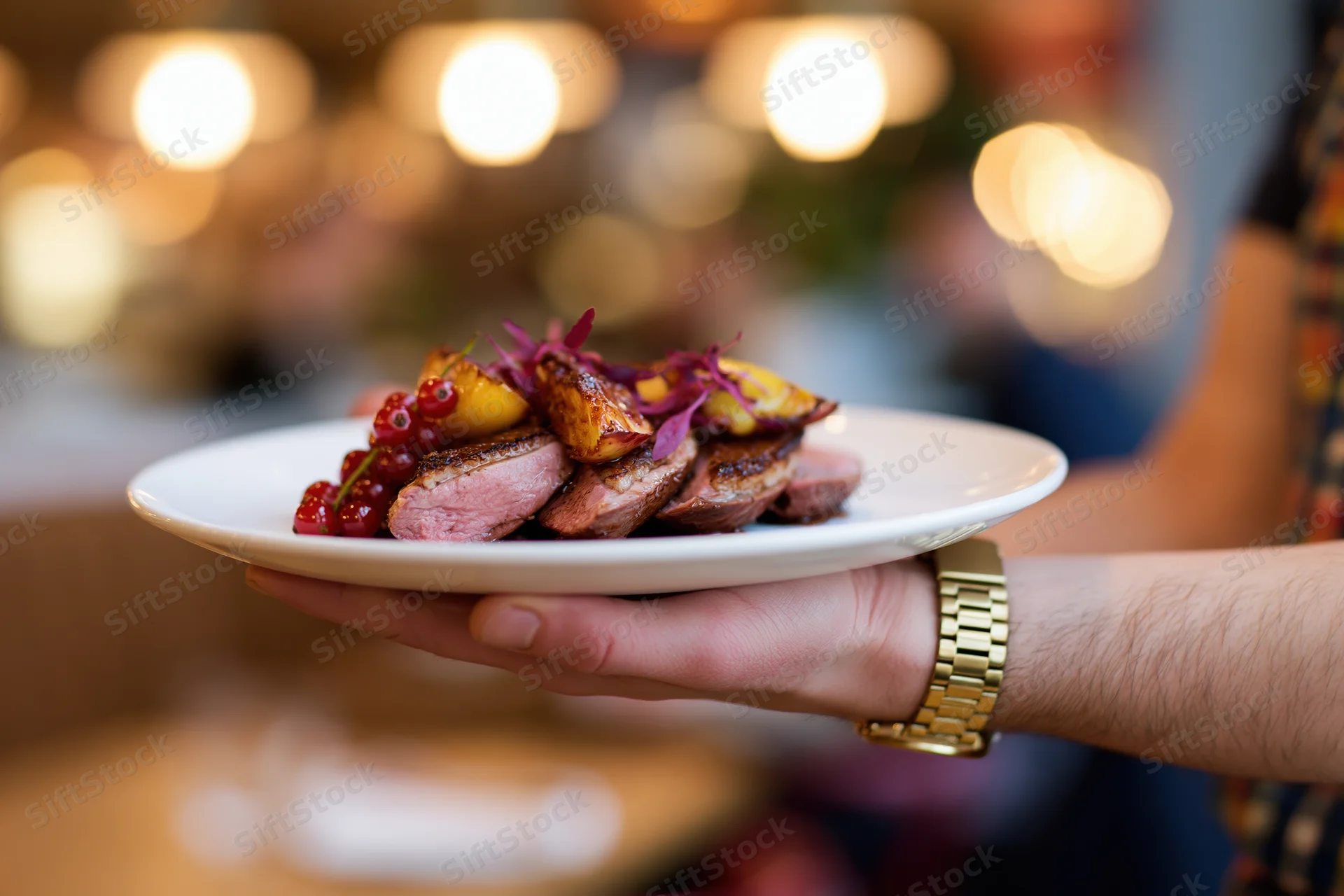 Hand holding a plated seared duck breast with vegetables and red currants in a restaurant setting