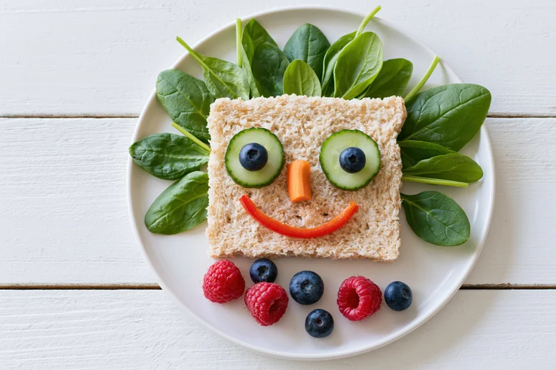 Kids sandwich decorated as a smiley face on a white plate with spinach and berries
