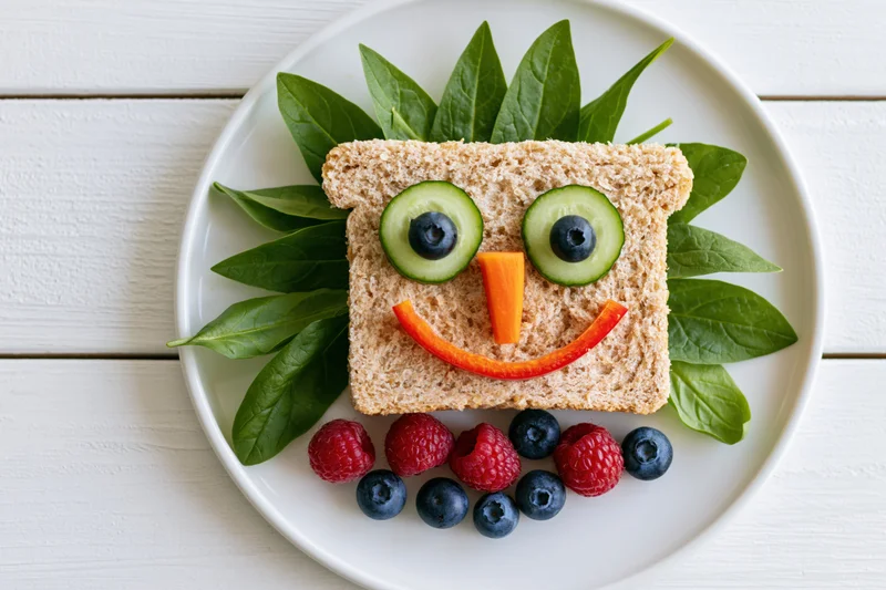 Kids sandwich decorated as a smiley face on a white plate with vegetables and berries