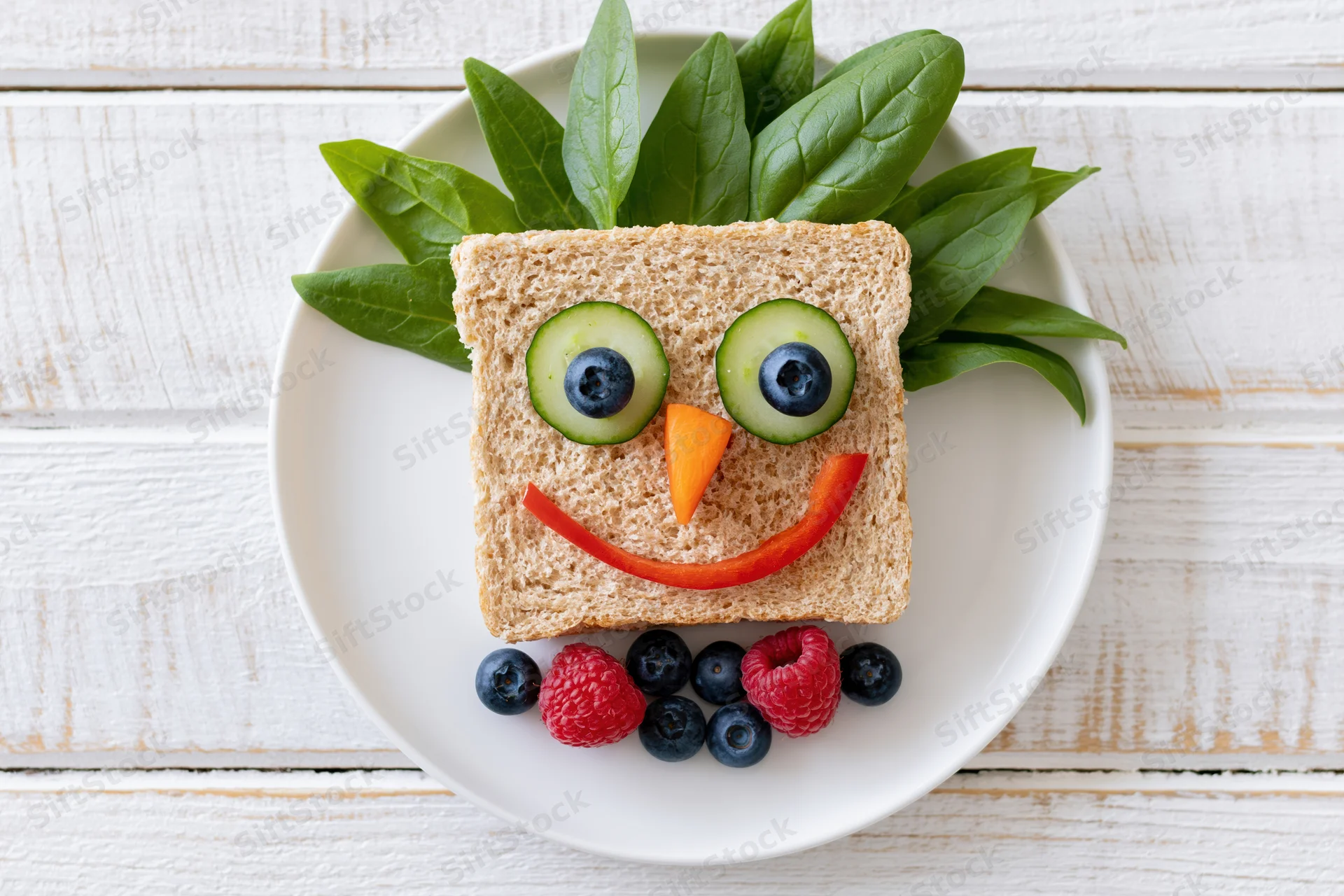 Kids sandwich decorated as a smiley face on a white plate with vegetables and berries