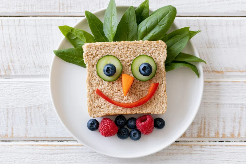 Kids sandwich decorated as a smiley face on a white plate with vegetables and berries