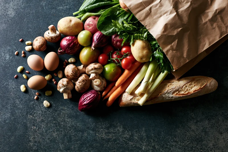 Kraft paper grocery bag tipped on its side spilling fresh vegetables fruits bread eggs and beans onto dark countertop