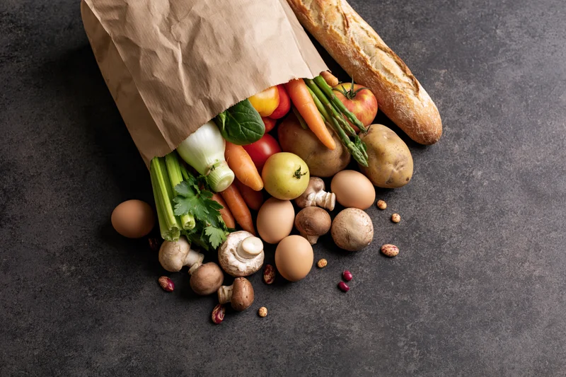 Kraft paper grocery bag tipped over spilling fresh vegetables, bread, eggs and mushrooms on dark surface