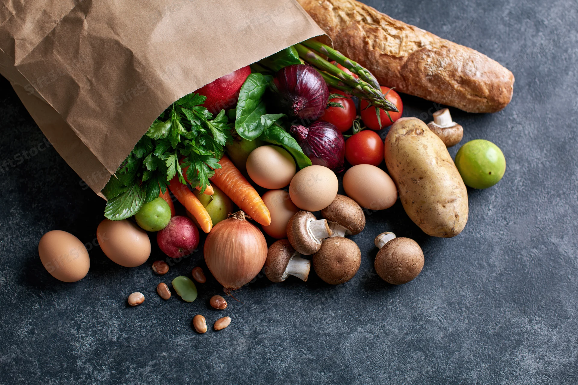 Kraft paper grocery bag tipped over spilling fresh vegetables fruits eggs bread and mushrooms on dark stone surface