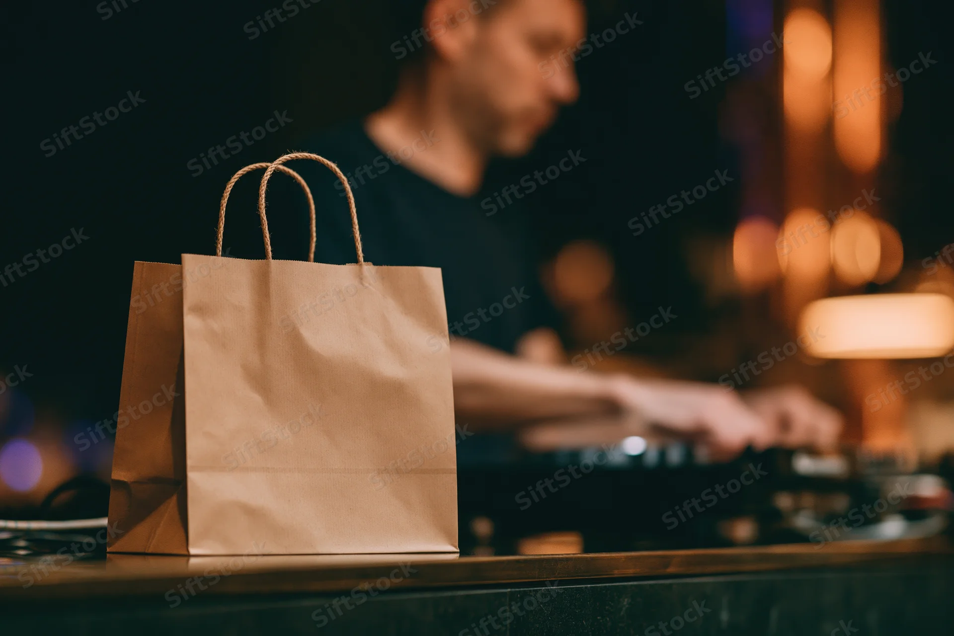 Kraft paper takeout bag with rope handles on a bar counter in front of a dj operating turntables at night