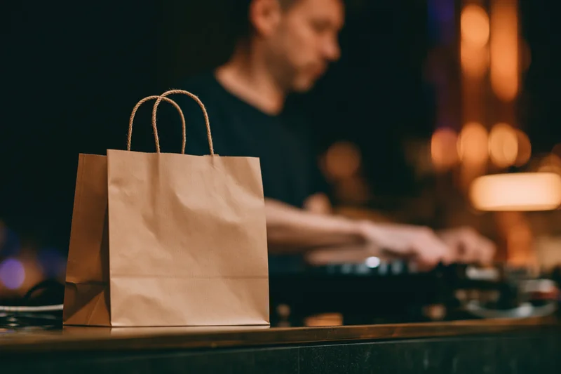 Kraft paper takeout bag with rope handles on a bar counter in front of a dj operating turntables at night