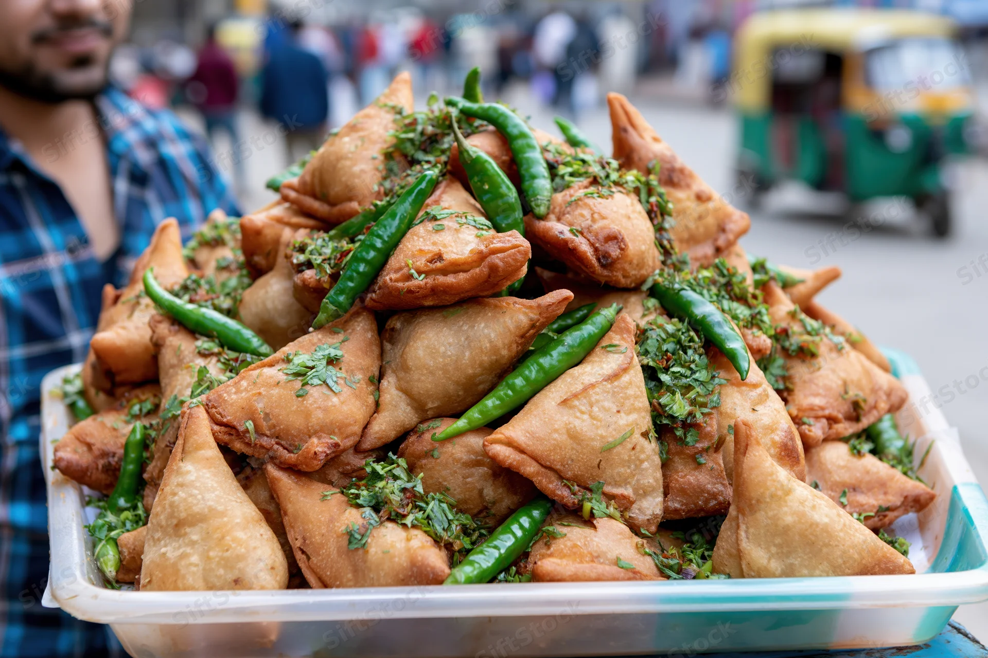 Pile of golden fried samosas topped with green chilies and cilantro on a plastic tray held by a street vendor in an urban market setting