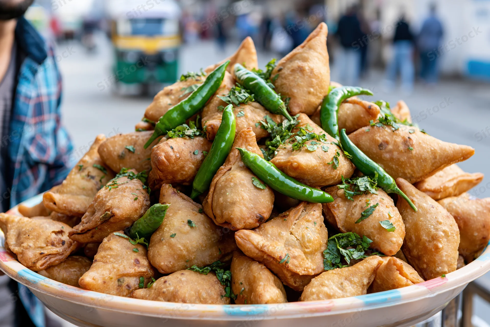 Golden fried samosas piled high in a bowl topped with green chilies and cilantro at a street market