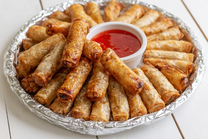 Fried lumpia springrolls piled on a foil-covered tray with red dipping sauce on a white wooden table