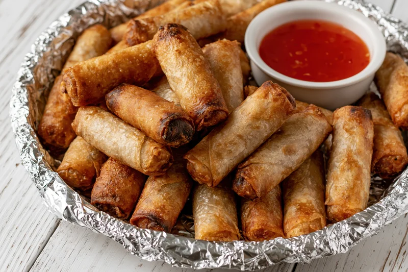 Fried lumpia springrolls piled on a foil-lined tray with red dipping sauce on a wooden table