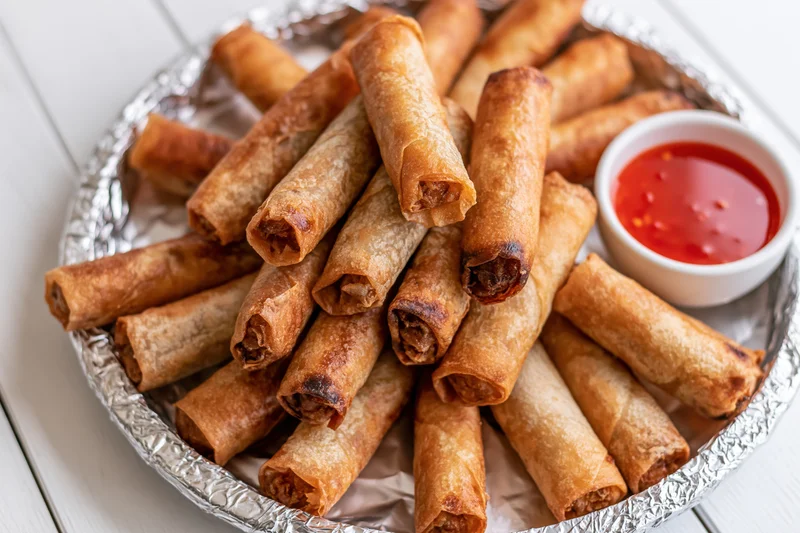 Fried lumpia springrolls piled on a foil-covered tray with sweet chili sauce on the side