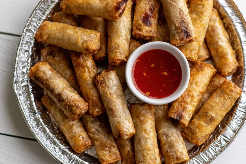 Fried lumpia springrolls piled on a foil tray with red dipping sauce in the center