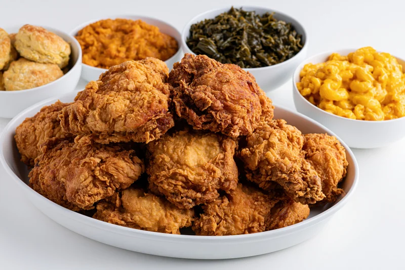 Golden-brown fried chicken piled high on a white oval platter with side dishes of macaroni and cheese, collard greens, sweet potato, and biscuits