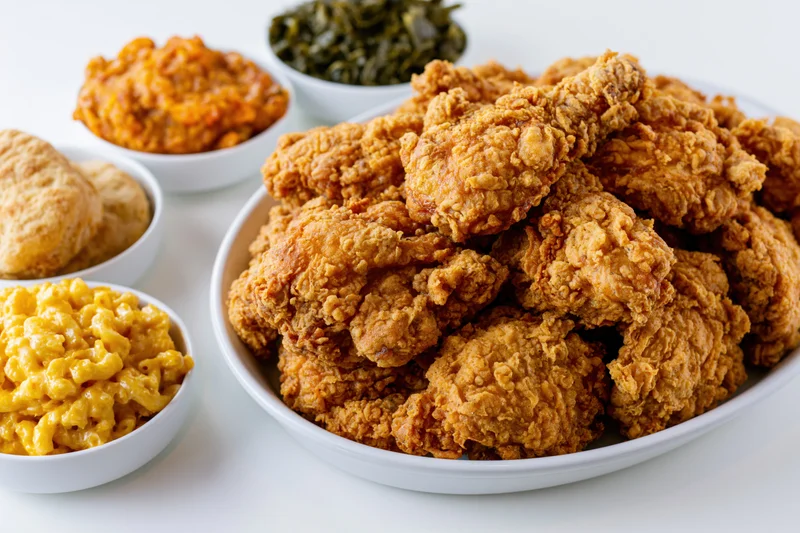 Golden-brown fried chicken piled high on a white oval platter with side dishes of macaroni and cheese, biscuits, collard greens, and sauce arranged around it on a white surface